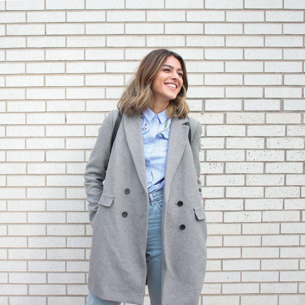 femme portant une chemise bleues un jean et un manteau long gris devant un mur en brique blanc