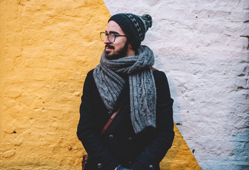 homme portant un bonnet un manteau foncé et une écharpe grise devant un mur blanc et jaune