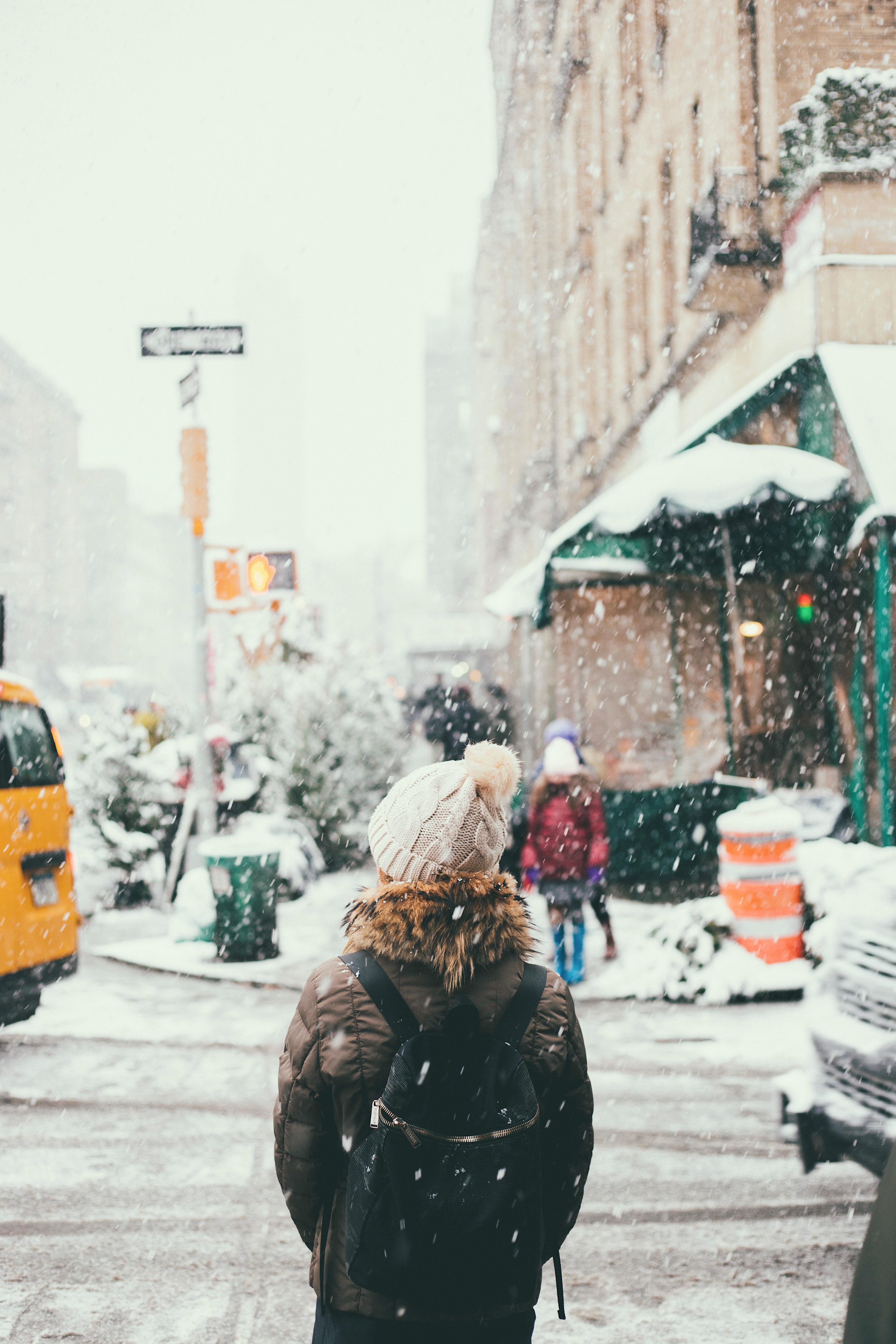 femme se tenant dans la rue en hiver sous la neige portant un bonnet un manteau kaki avec de la fourrure au niveau de la capuche avc un sac à dos foncé